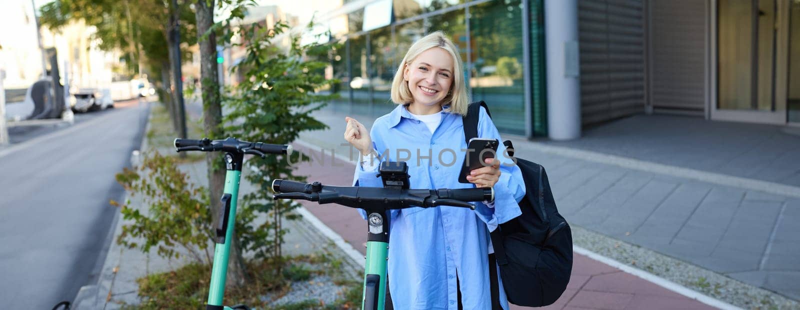 Beautiful blond girl, scans QR code on mobile phone, stands near electric street scooter to rent it, requesting a ride, smiling and looking happy.