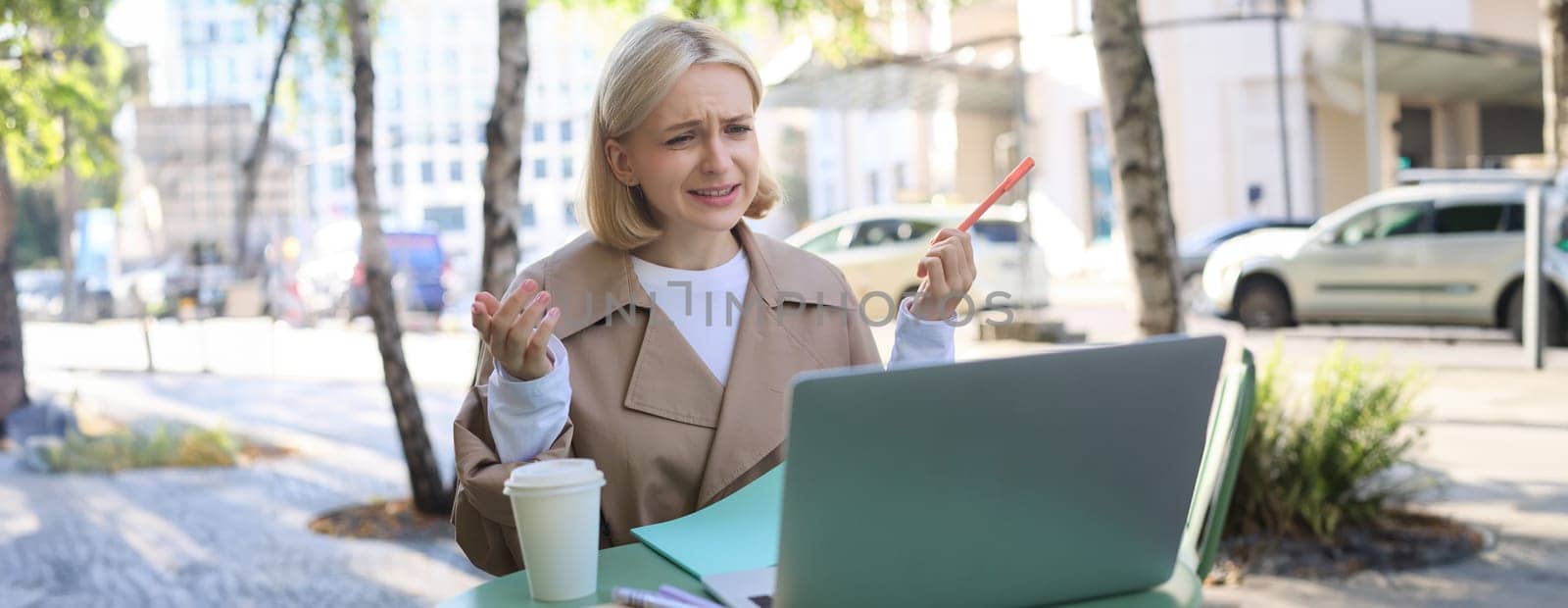 Portrait of young female student, modern woman in outdoor coffee shop, street cafe, talking to someone via laptop, video chatting, communicating, working remotely online.