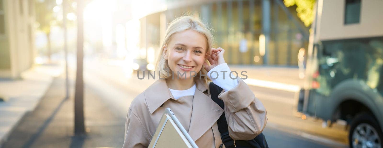 Young blonde woman, college student on street, holding journals and notebook, carry her homework material, going to university, smiling and looking happy.
