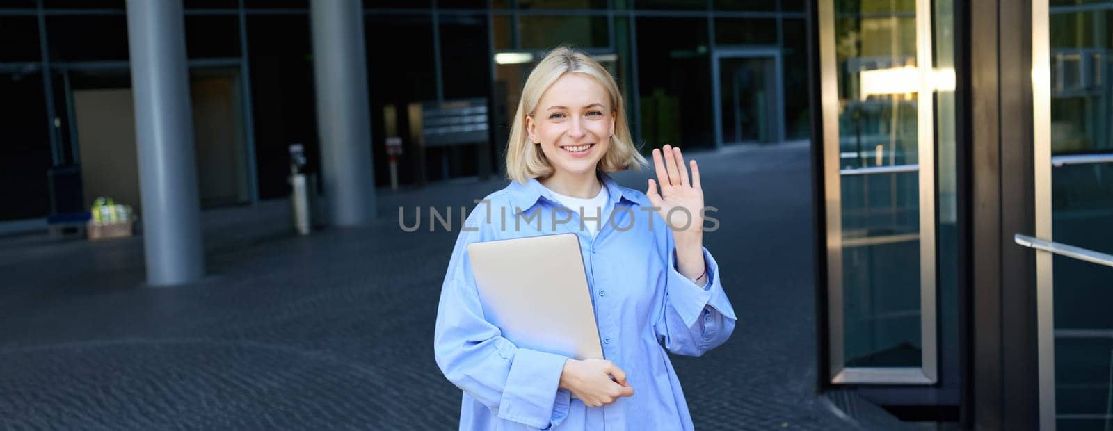 Friendly smiling student, blond girl with notebooks, tutor waving hand at camera, saying hello, standing near office building outdoors.