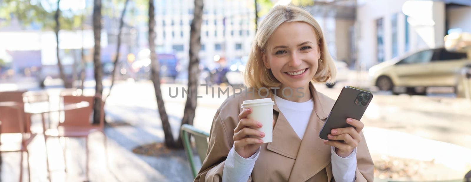 Portrait of cute blond woman with smartphone, drinking chai, coffee at favourite place in city centre, sitting outdoors, holding mobile phone by Benzoix