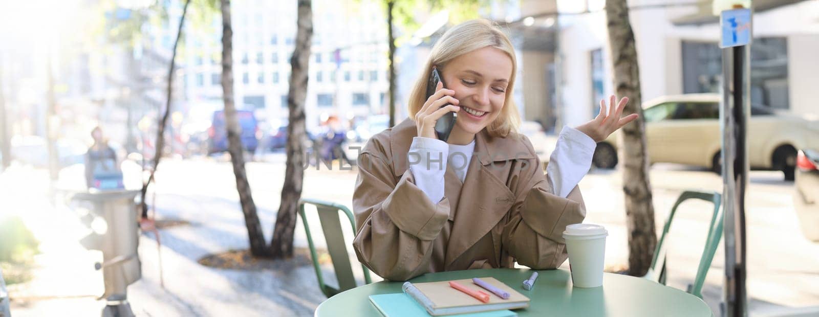 Outgoing young happy woman, sitting in cafe outdoors, talking on mobile phone, smiling, spending time outside in city centre, chatting. Lifestyle and people concept