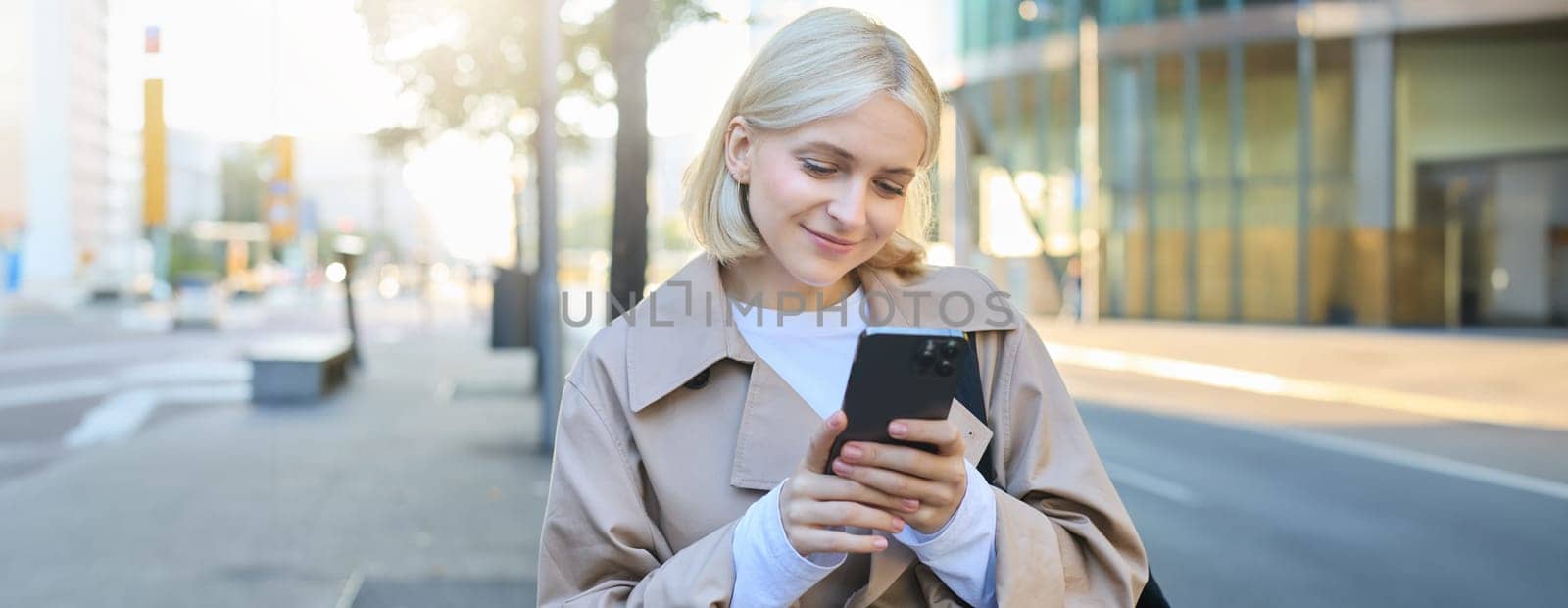 Image of young modern woman, student walking on street and looking at her mobile phone, smiling and looking happy by Benzoix
