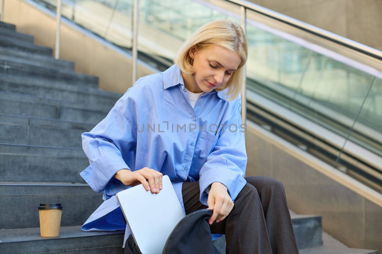 Young modern woman sits on stairs outdoors, drinks coffee, packs her laptop inside backpack. Lifestyle and student concept