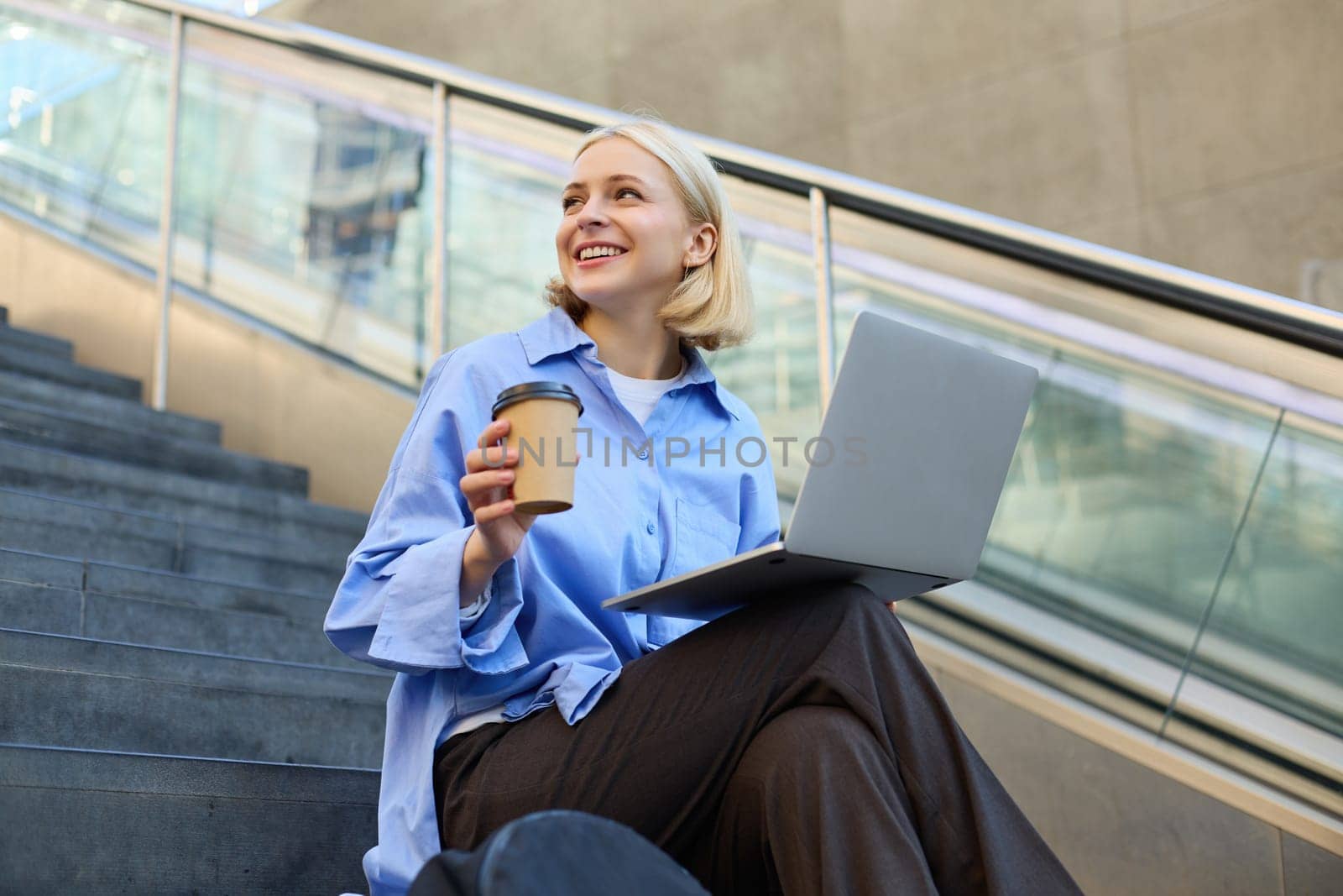 Young woman, student in blue shirt, working on laptop, sitting outdoors on street stairs, working on project online, connects to public wifi, elearning.