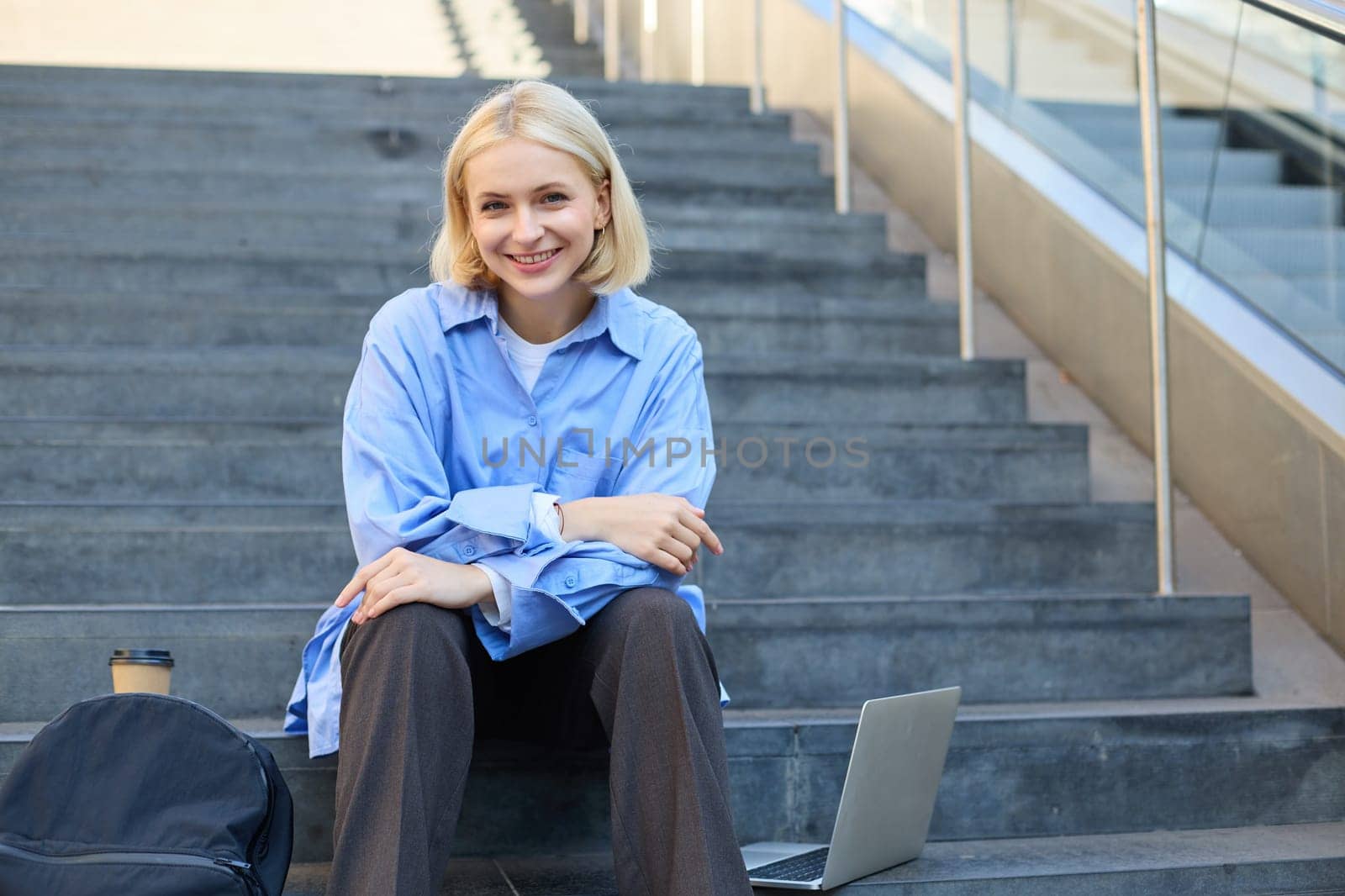 Urban style portrait of young woman, student sitting on city street stairs, with backpack, cup of coffee and laptop, looking happy and upbeat, smiling by Benzoix