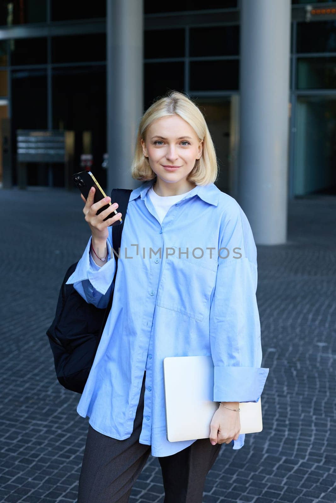 Vertical portrait of young office manager, woman in blue collar shirt and backpack, holding laptop and smartphone, waiting near business building on street, smiling at camera by Benzoix