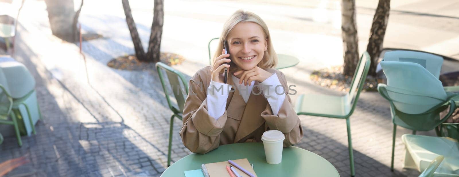 Happy blond female model, sitting outdoors in cafe, answer phone call, having conversation on smartphone, laughing and smiling. Lifestyle and people concept