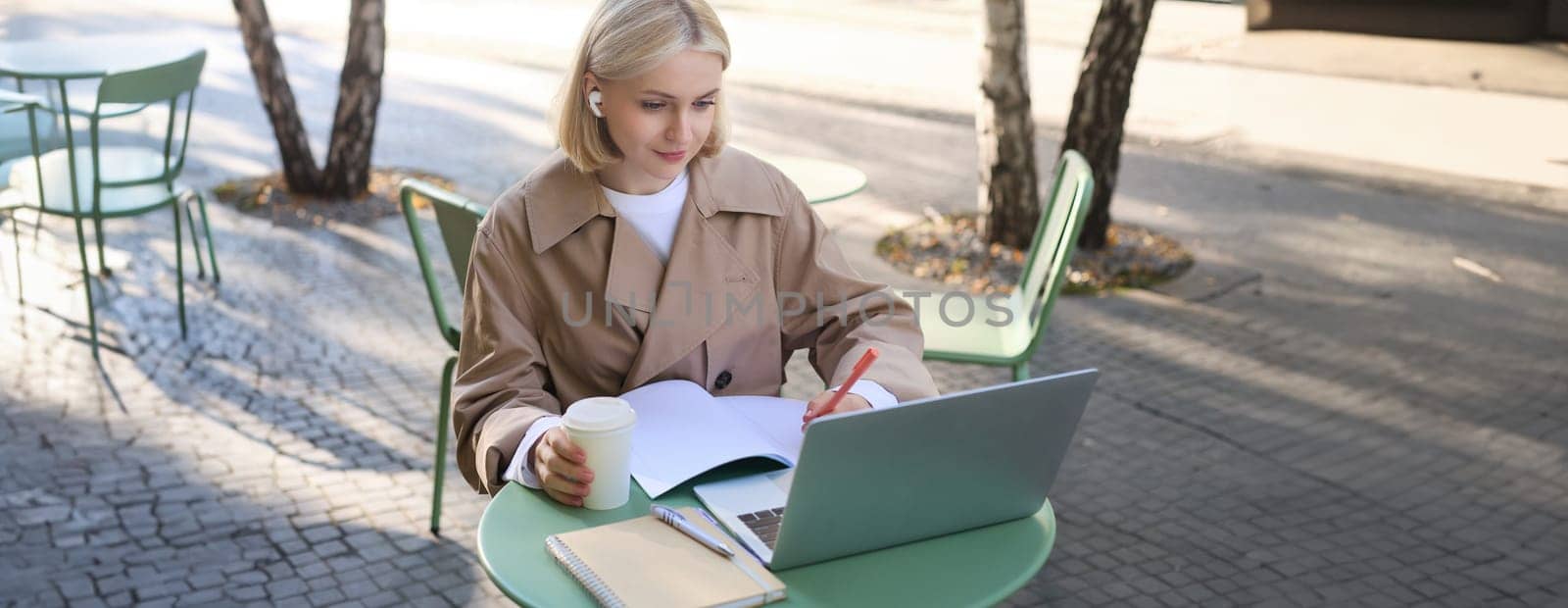 Portrait of beautiful blond woman, wearing wireless headphones, using laptop, studying in outdoor coffee shop, making notes, working on project.