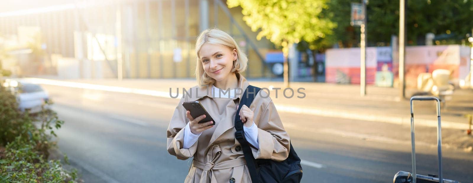 Smiling, beautiful young woman with backpack, holding smartphone, standing on street, using mobile phone app.
