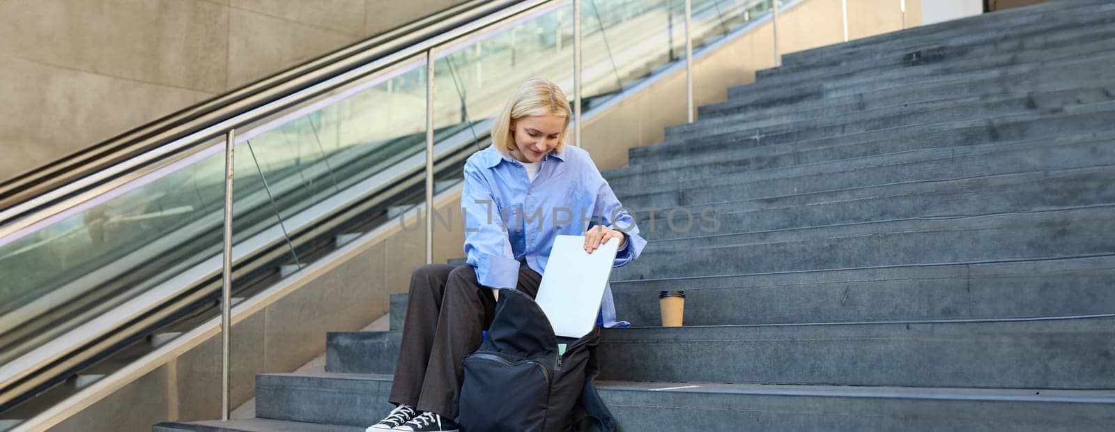 Portrait of young modern woman, student freelancer, sitting on public stairs outdoors, putting away her laptop, packing backpack, drinking coffee outside.