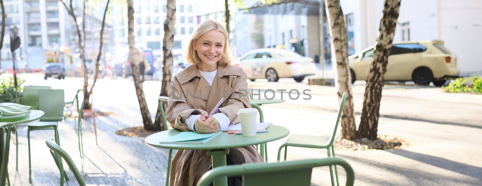 Portrait of young concentrated woman, doing homework in a cafe, writing in notebook, working with documents and drinking coffee outdoors.