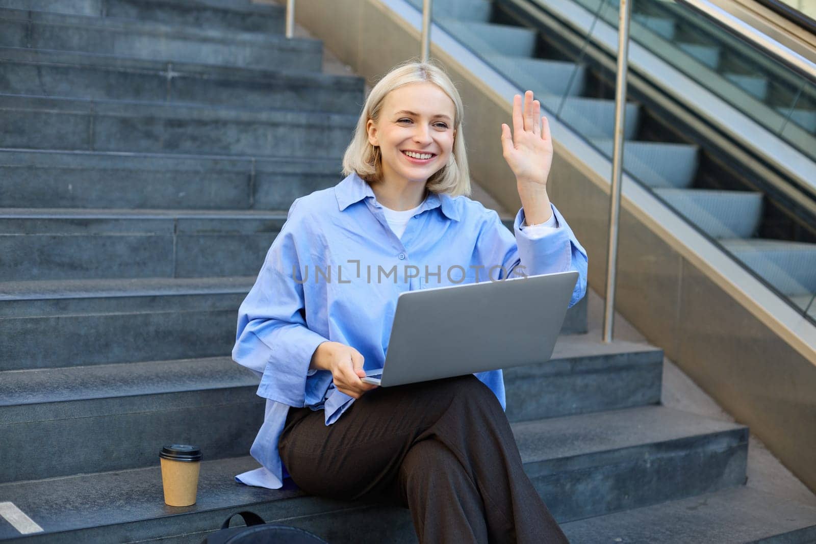 Happy, friendly young woman sitting with laptop on street stairs, waving hand to say hello, working remotely, elearning, connecting to public wifi by Benzoix