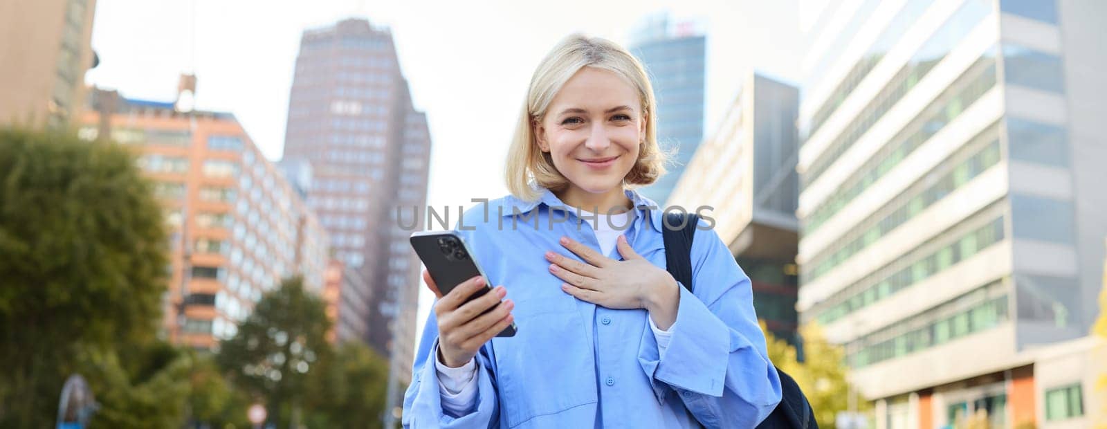 Image of young professional, office manager woman with backpack and smartphone, posing on streets of busy city, holding hand on chest, smiling and looking excited at camera by Benzoix