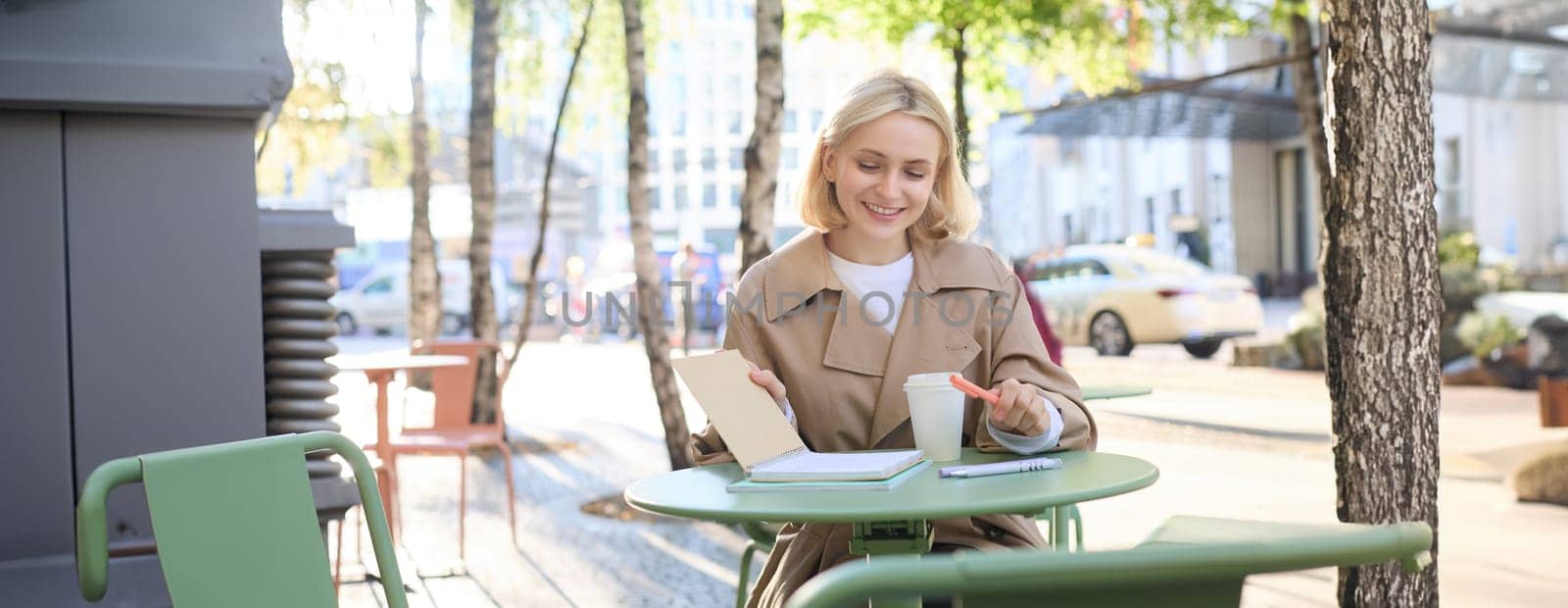 Portrait of stylish modern woman, sitting in an outdoor cafe, smiling and drinking coffee from takeaway cup, wearing trench coat.