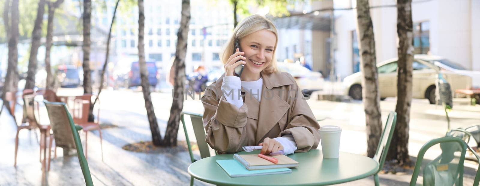 Lifestyle and urban people concept. Young smiling girl, student sitting in cafe outdoors, talking on mobile phone, chatting with friend or coworker over the telephone, studying or doing homework by Benzoix