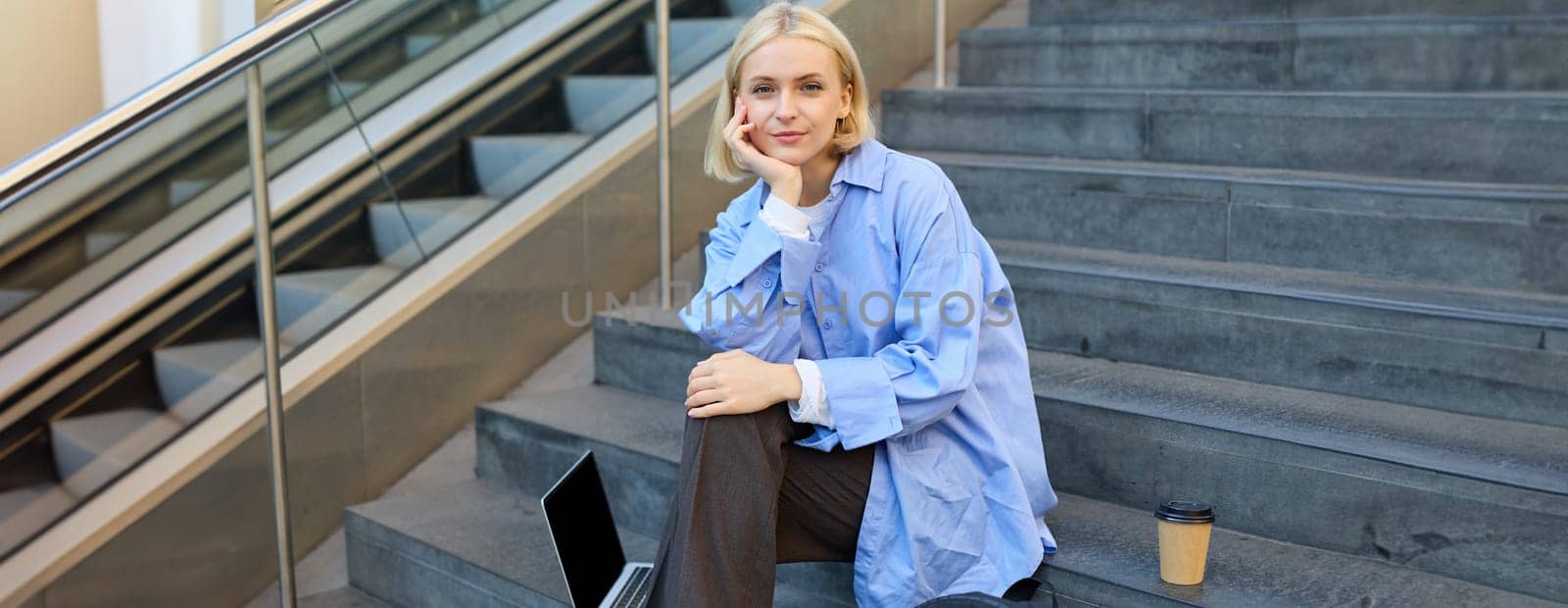 Portrait of young modern woman, student or freelancer, sitting on outdoor stairs, resting in city, has laptop and coffee, looking at camera with confident, smiling face by Benzoix