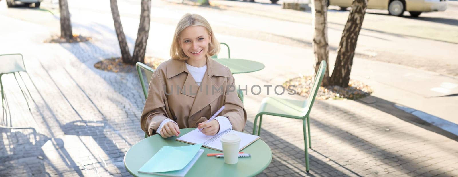 Image of young blonde woman in stylish trench, sitting in outdoor cafe, drinking coffee and working on documents, studying, doing homework in her notebook, smiling at camera by Benzoix