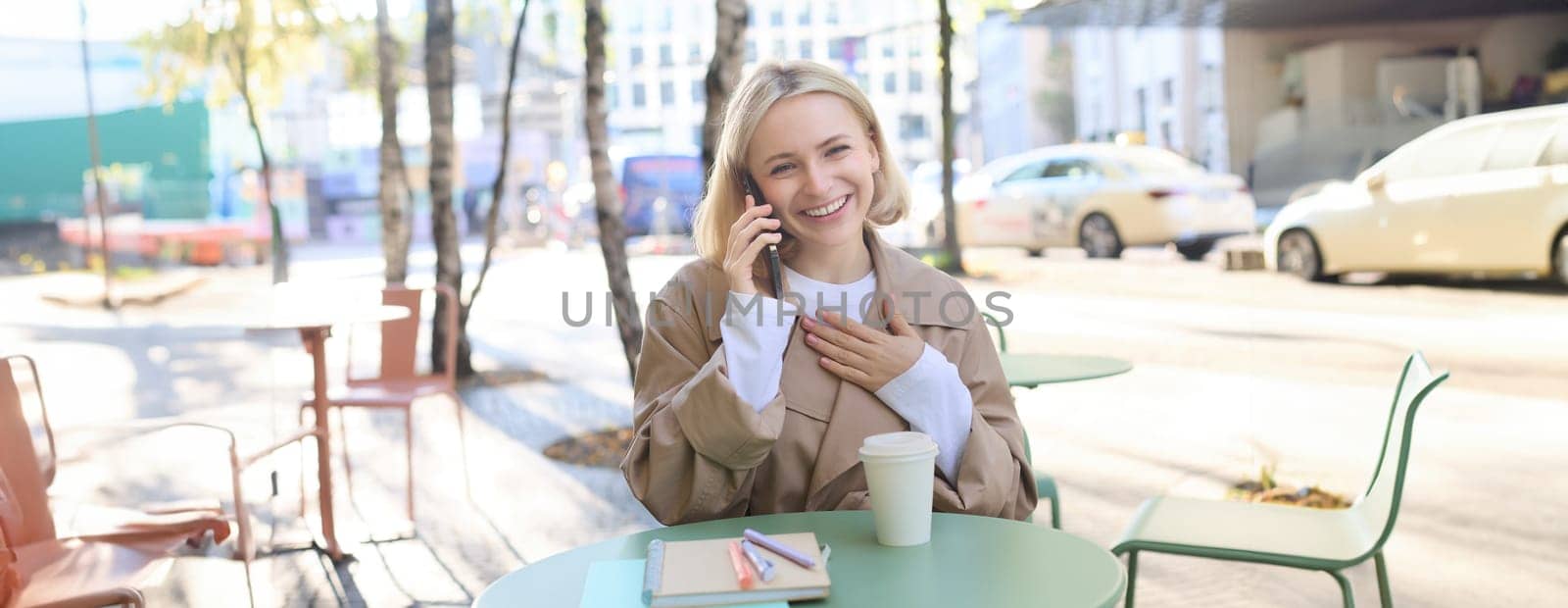 Happy blond female model, sitting outdoors in cafe, answer phone call, having conversation on smartphone, laughing and smiling by Benzoix