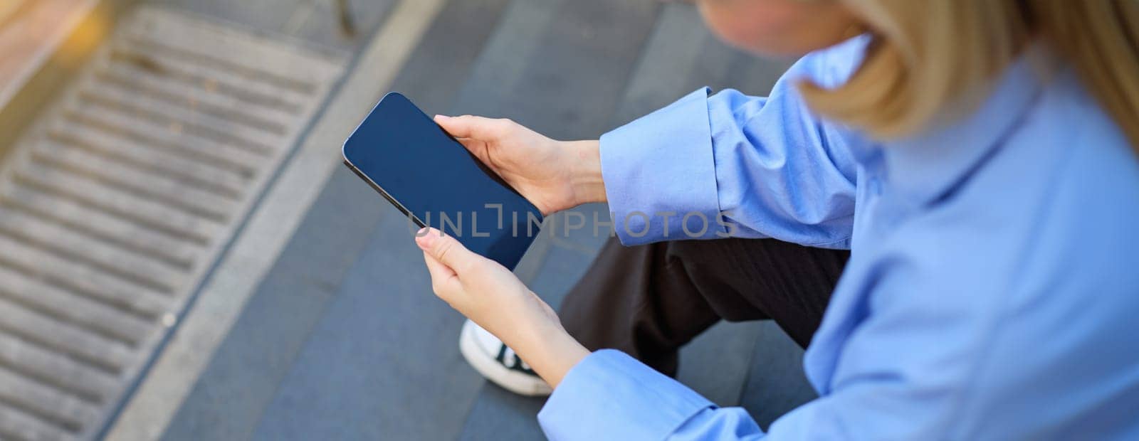 Shot from behind, young woman sitting on stairs on street, holding mobile phone in both hands, screen is dark and blank.