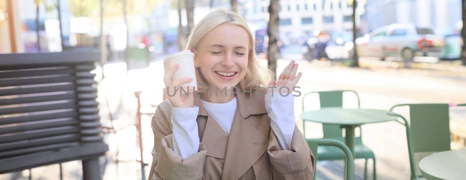 Portrait of cheerful young blond woman, enjoying spending time in outdoor cafe, drinking coffee and laughing, having casual meet up in street restaurant by Benzoix