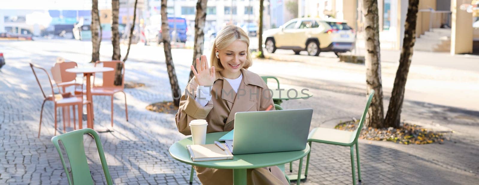 Cheerful girl with laptop, saying hello, connects to online meeting, waving hand at camera, chatting with someone via internet, sitting in outdoor coffee shop by Benzoix
