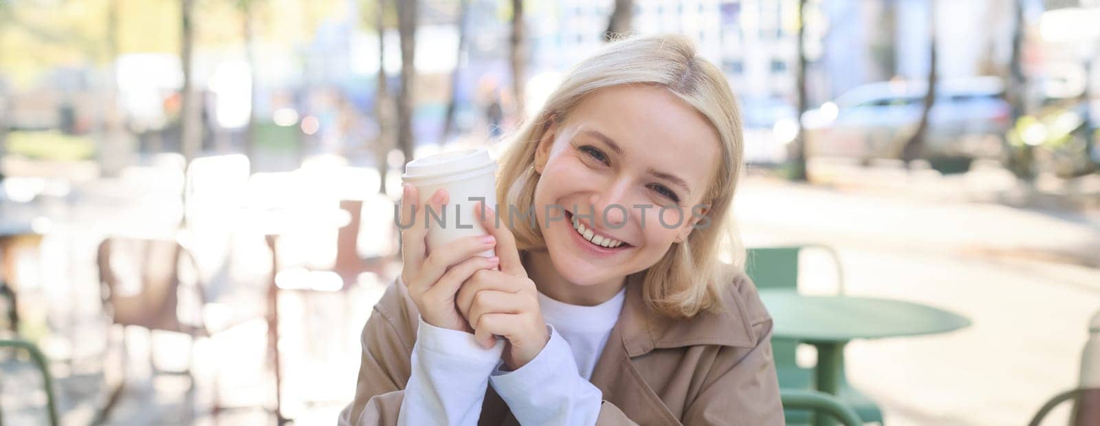Close up portrait of smiling blonde woman, university student, holding cup of coffee, warming up her hands on chilly autumn day, sitting in outdoor cafe, laughing at camera.