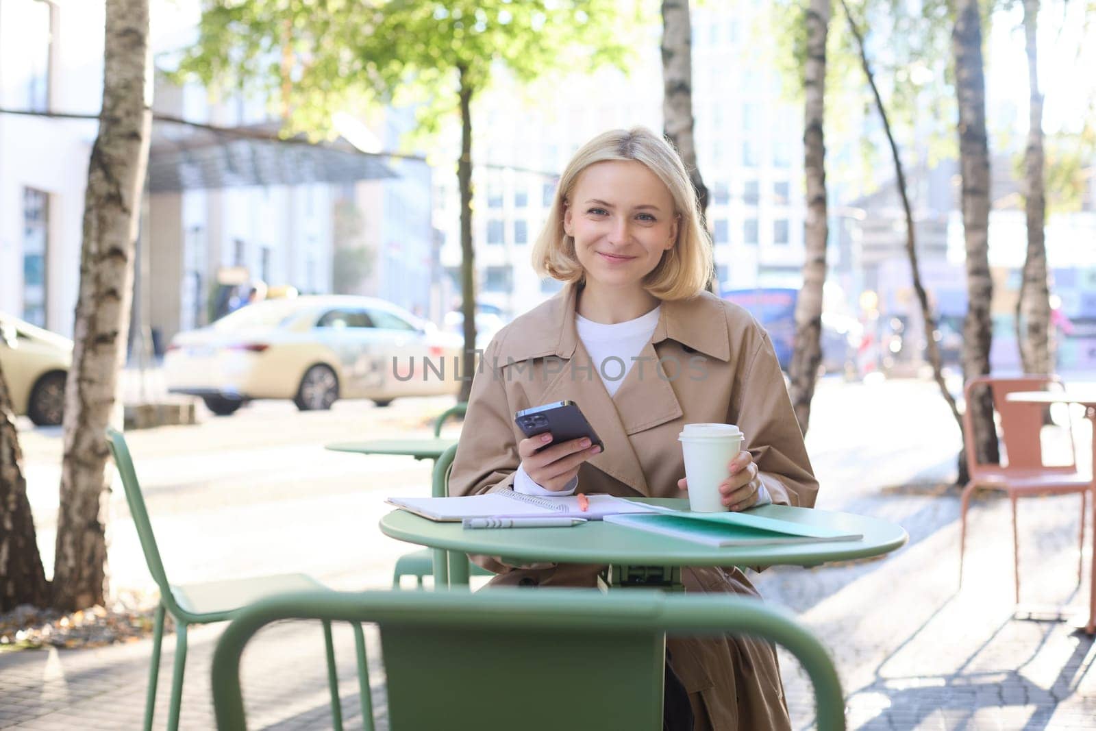 Portrait of young woman studying, sitting in outdoor cafe with smartphone and journals, doing homework, wearing trench coat on sunny day.