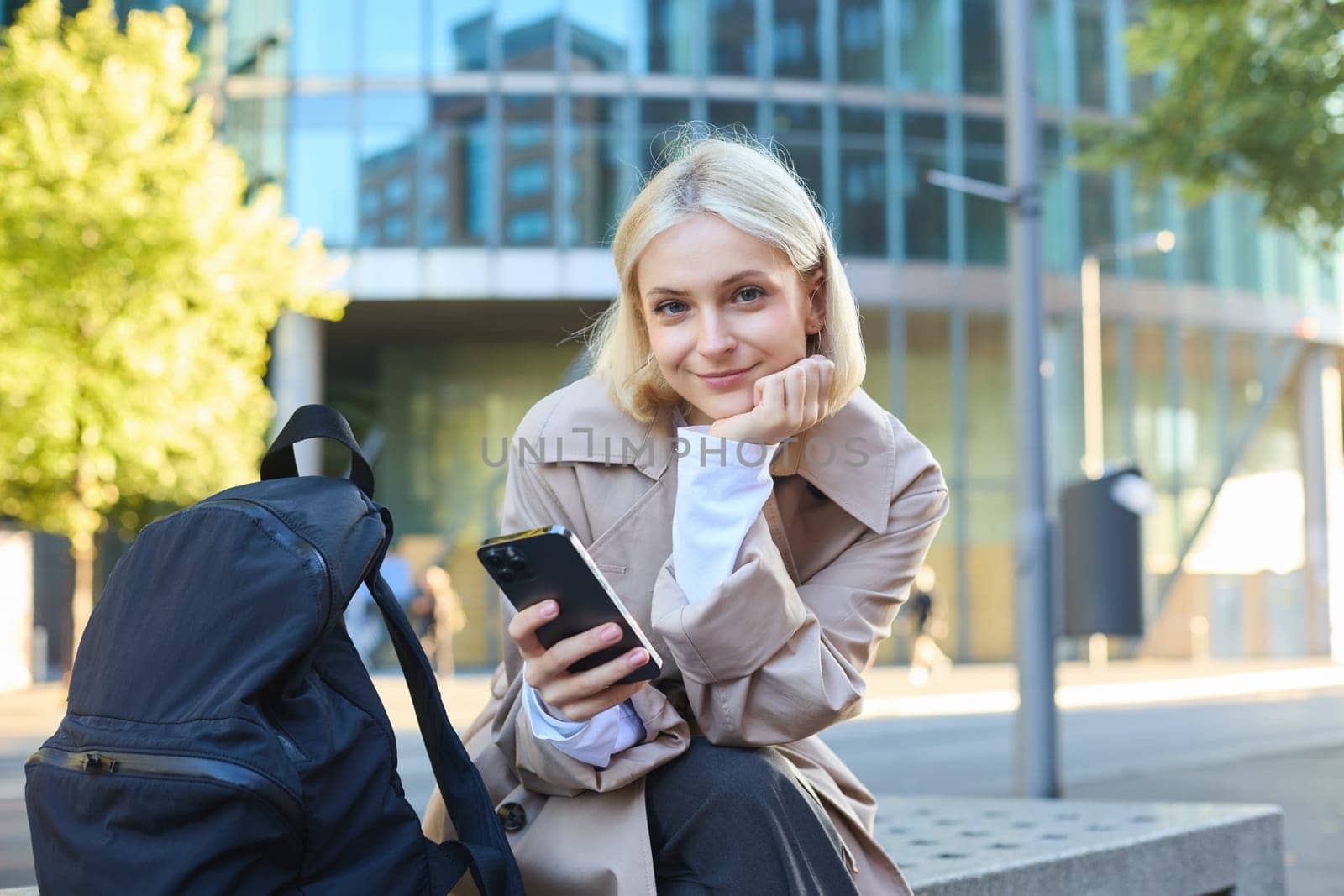 Portrait of young blonde woman, waiting for someone on street bench, sitting with smartphone, using mobile phone app by Benzoix