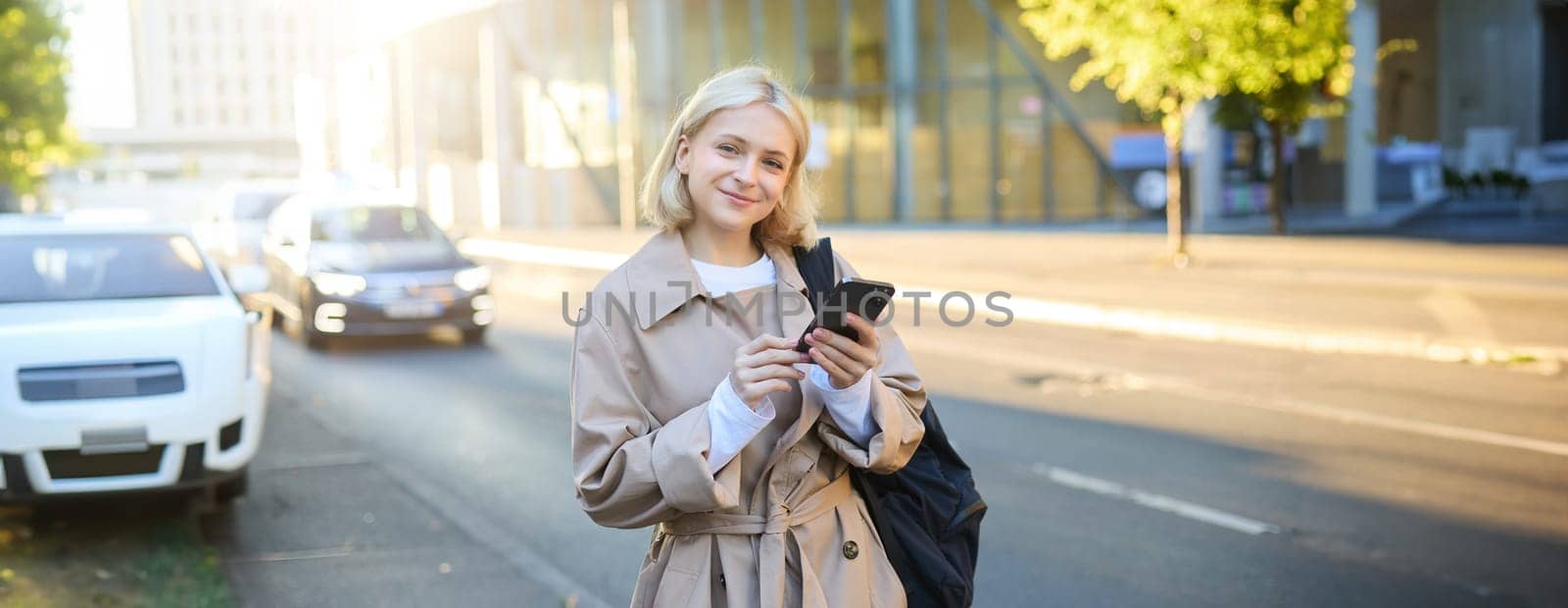Lifestyle portrait of young woman on street, standing near road with cars, carries backpack and holds mobile phone, requests a ride on smartphone app, smiling and looking happy at camera.