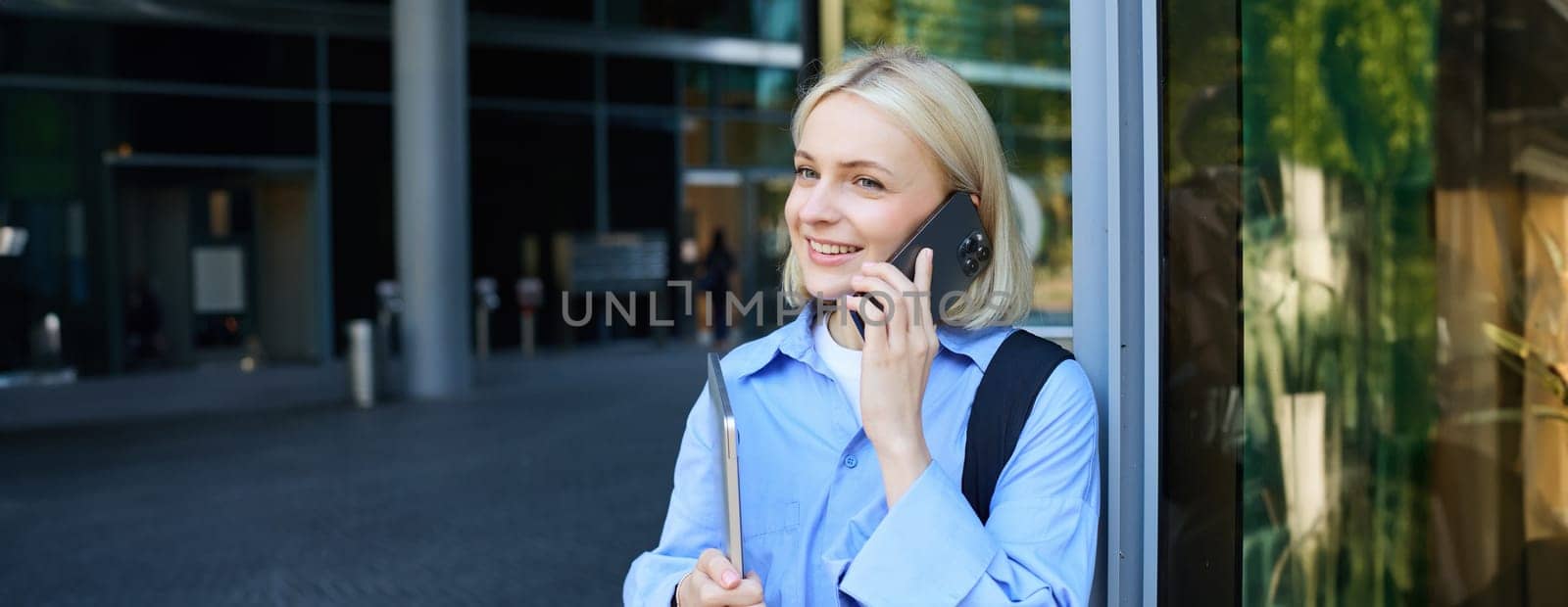 Portrait of young modern woman, office manager near building, standing outside with backpack, laptop, talking on mobile phone, chatting on smartphone. Lifestyle and communication concept