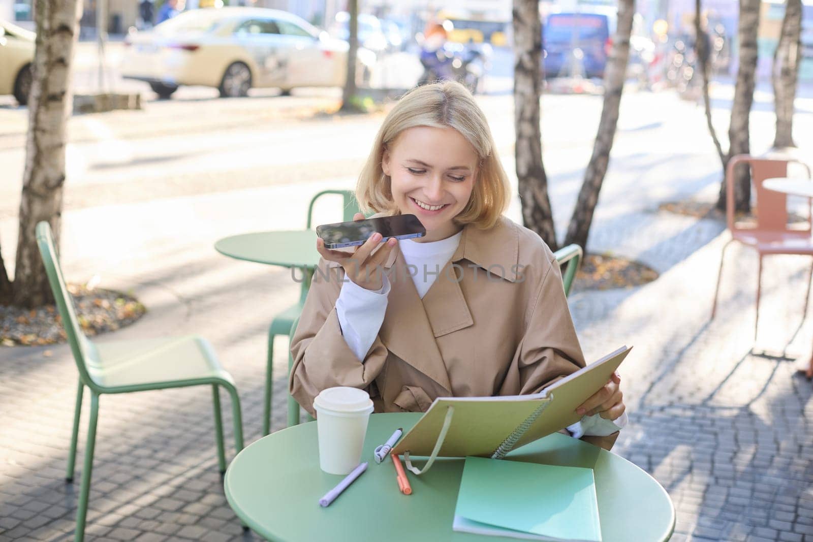 Lifestyle portrait of happy young woman, sitting in cafe and recording voice message, holding mobile phone near lips and talking into microphone, spending time in cafe, drinking coffee and chatting.