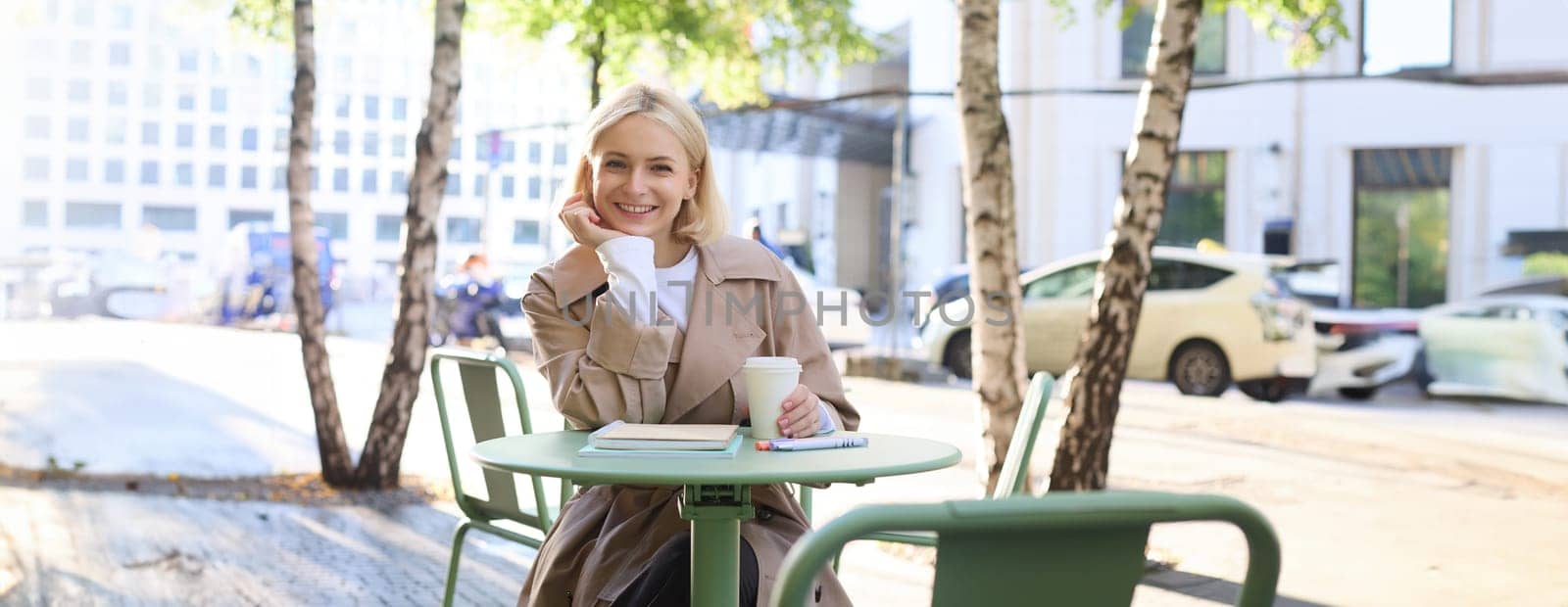 Urban lifestyle concept. Young beautiful woman sitting in outdoor cafe, contemplating city life, drinking coffee and smiling on street.