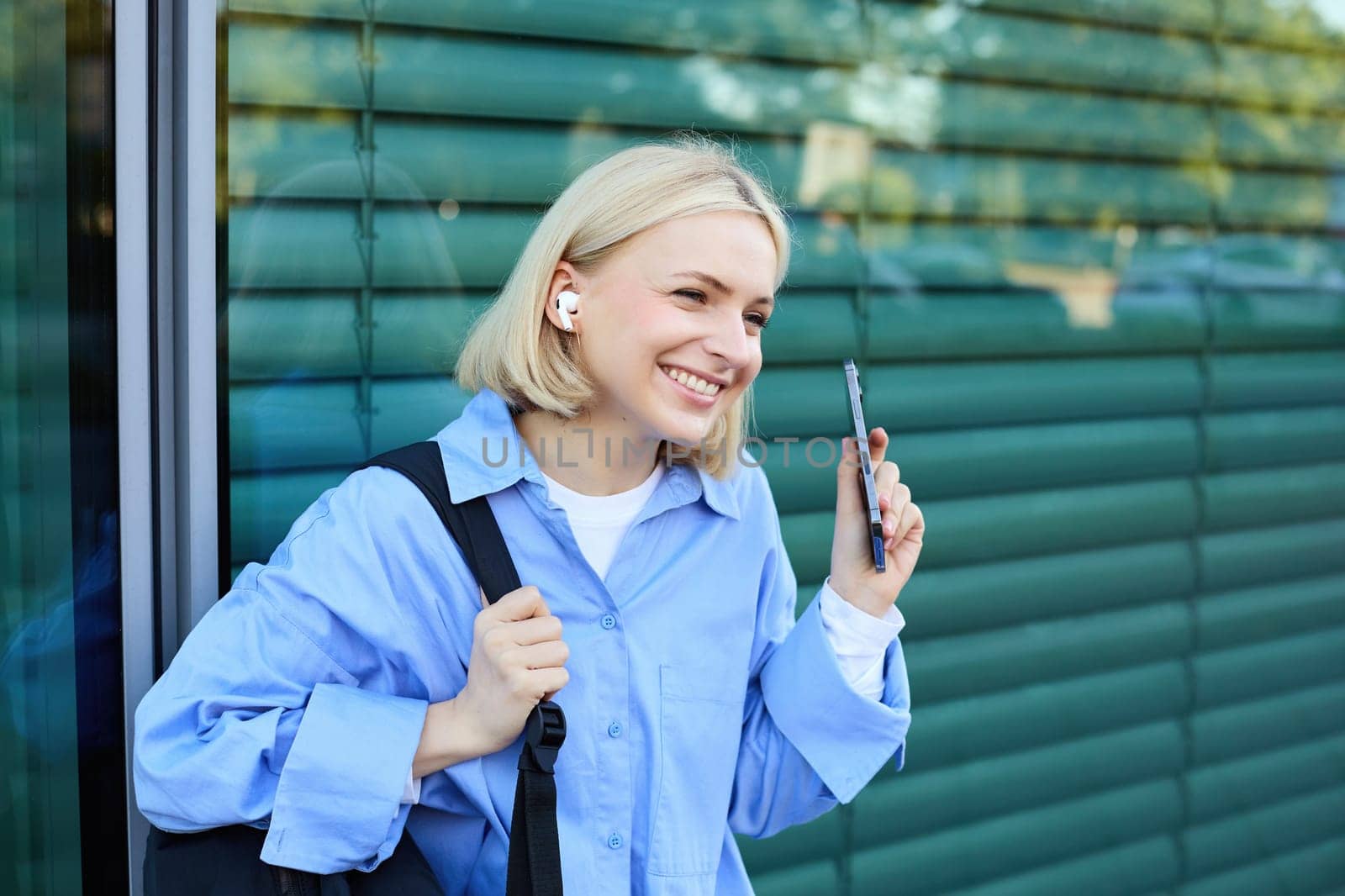Close up portrait of blond smiling female model, college student in earphones, listening to music on street, holding smartphone, waiting for someone outdoors.