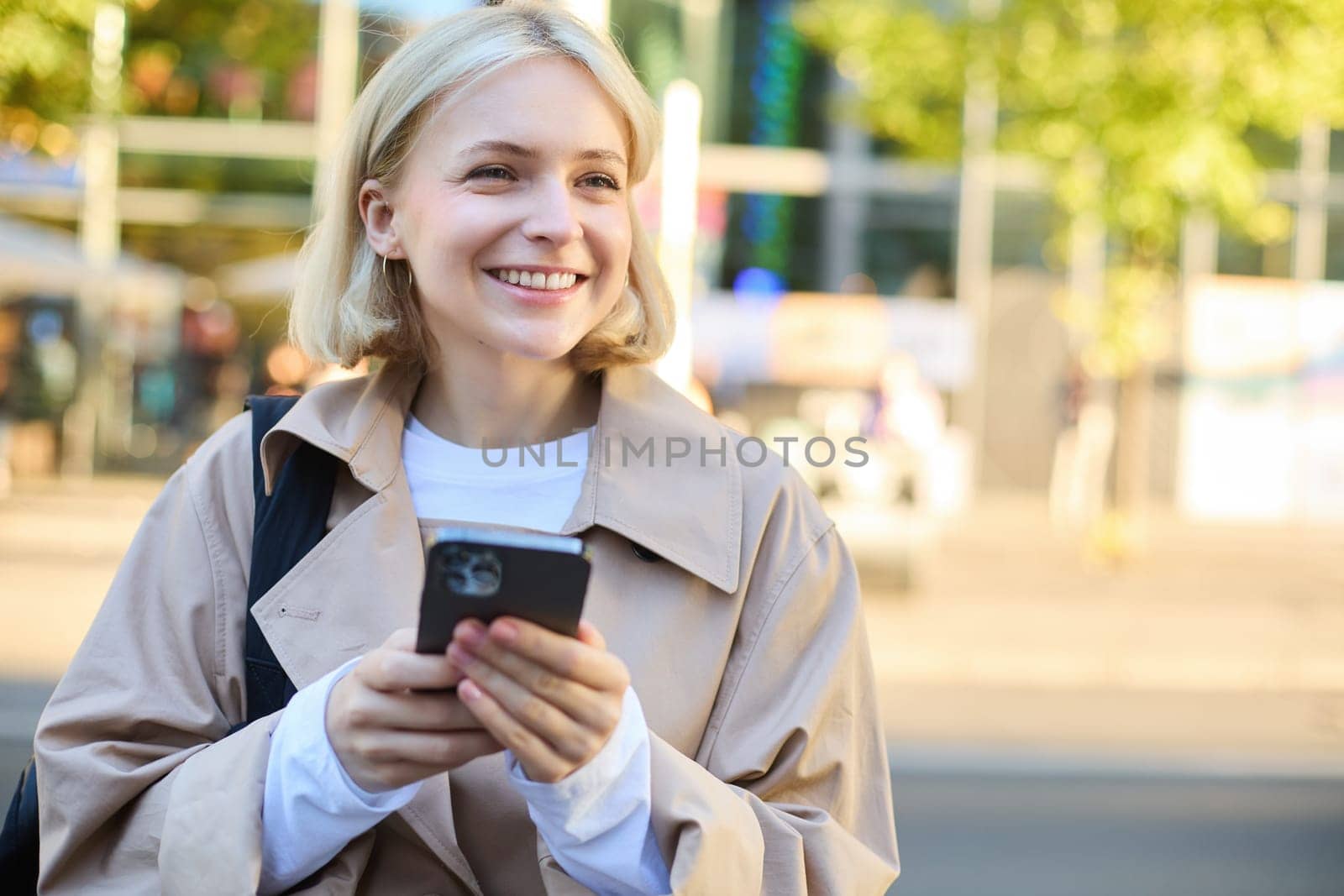 Carefree smiling woman standing on sunny street, using mobile phone, waiting for car or bus with smartphone, looking around.