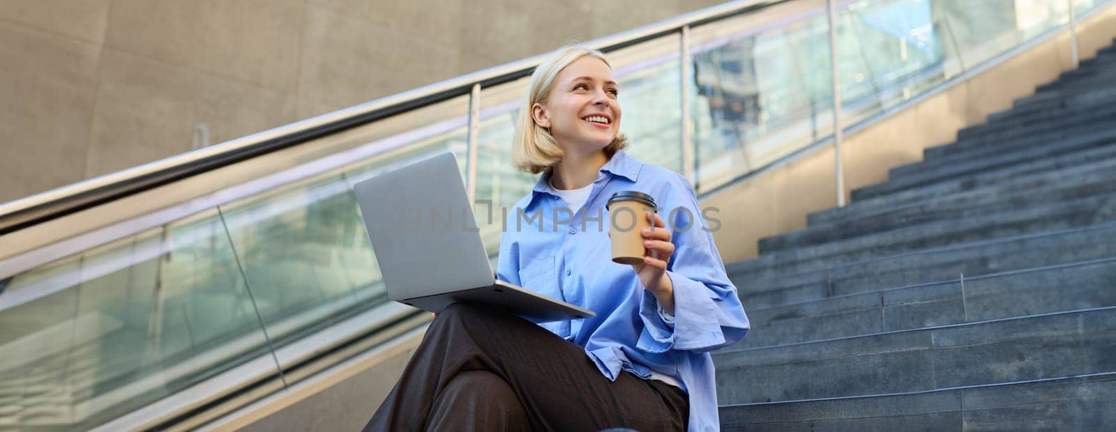 Young woman, student in blue shirt, working on laptop, sitting outdoors on street stairs, working on project online, connects to public wifi, elearning by Benzoix