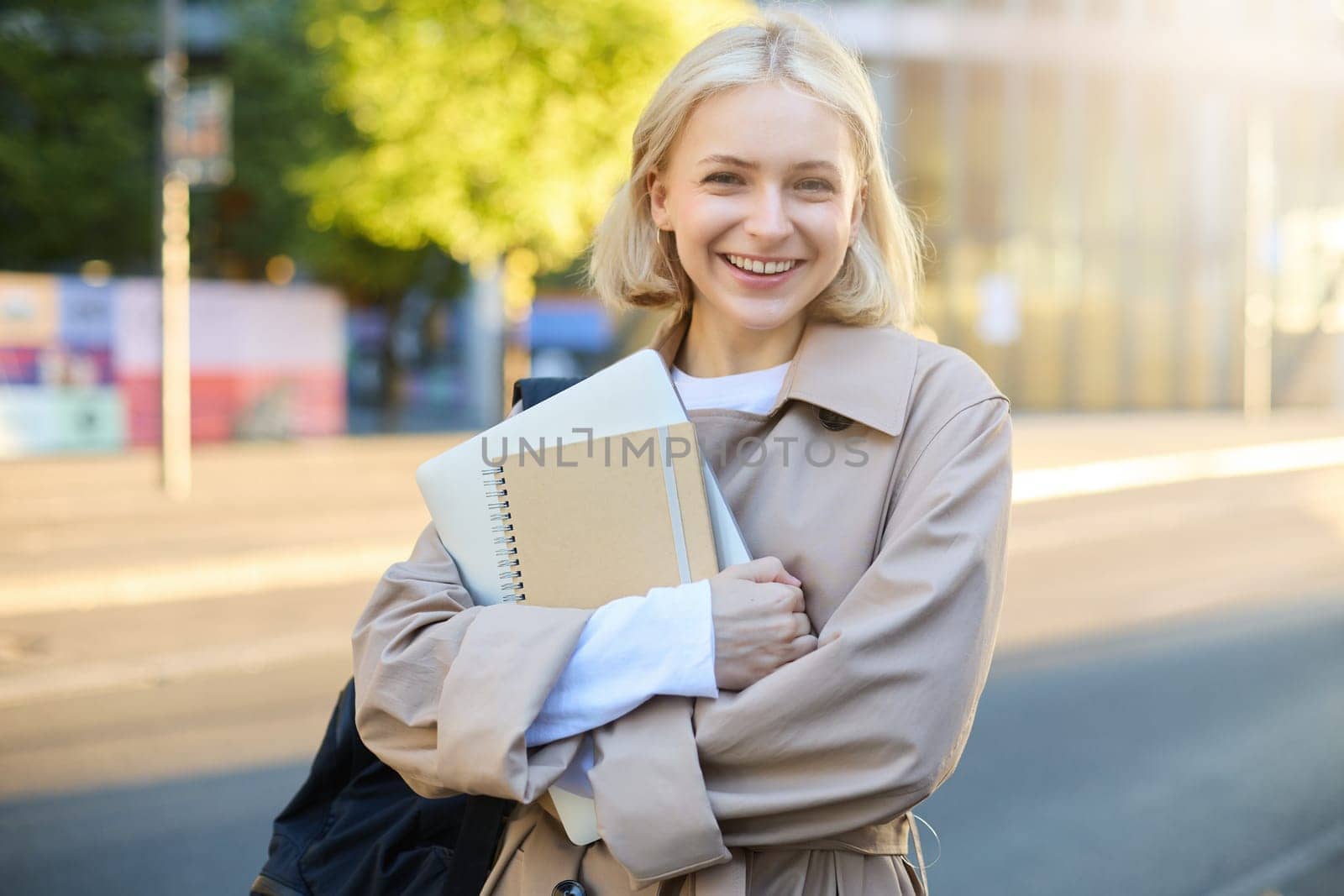 Portrait of beautiful blond girl, college student carries notebooks, holds backpack, smiles and looks happy at camera by Benzoix