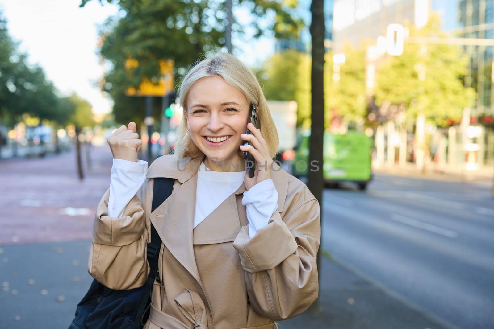 Close up portrait of beautiful young woman, blonge girl walking on street with mobile phone, chatting with friend, has happy face expression while talking over cellphone on her way.