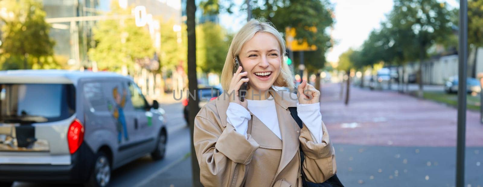 Portrait of cheerful girl talking on mobile phone, laughing while walking along the street, has backpack on shoulder. Lifestyle and people concept