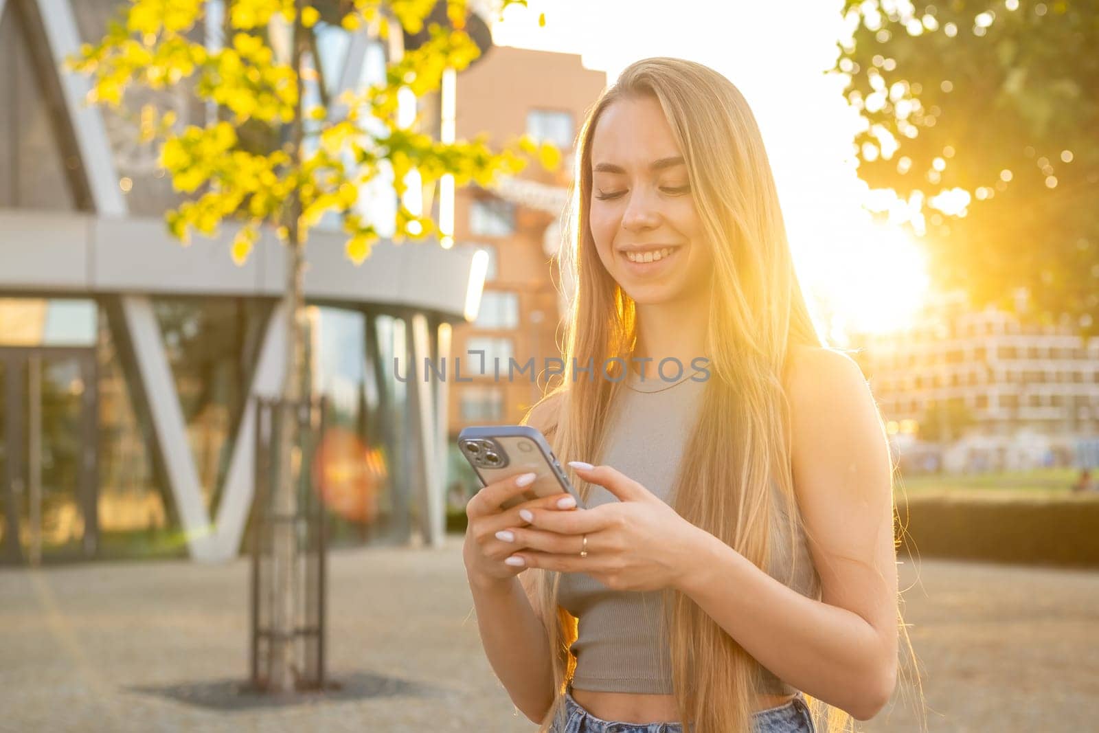 Happy young woman with long blonde hair holds a mobile phone standing in the street.