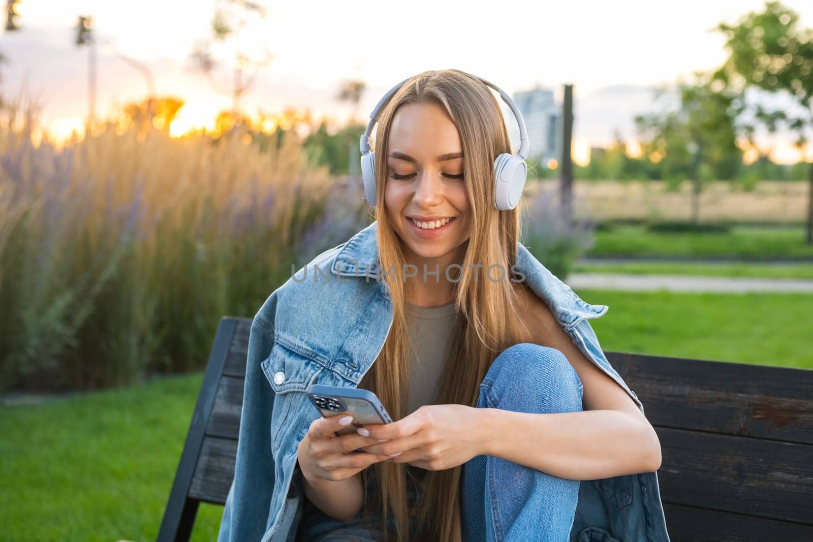 Young blonde woman in denim uses a mobile phone and listen to music sitting on the bench in the park