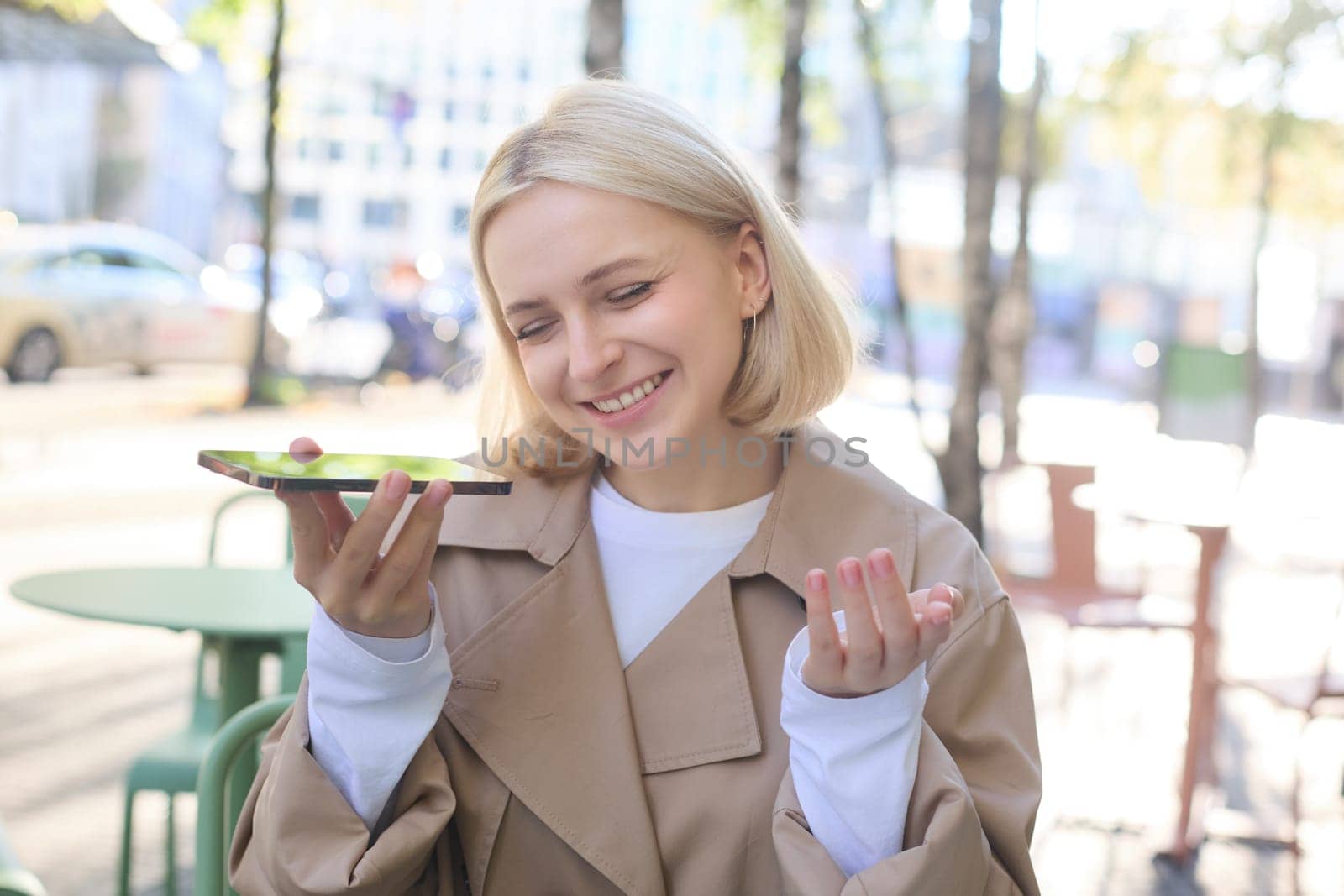 Close up portrait of beautiful young blond girl, records voice mail, holding smartphone near mouth and smiling, sitting in cafe outdoors.