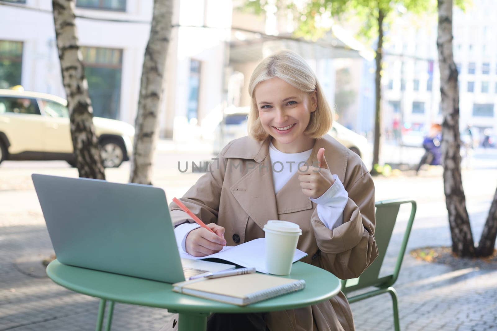 Portrait of beautiful blond female model, showing thumbs up, sitting with laptop and study material in coffee shop, gives approval. Lifestyle concept