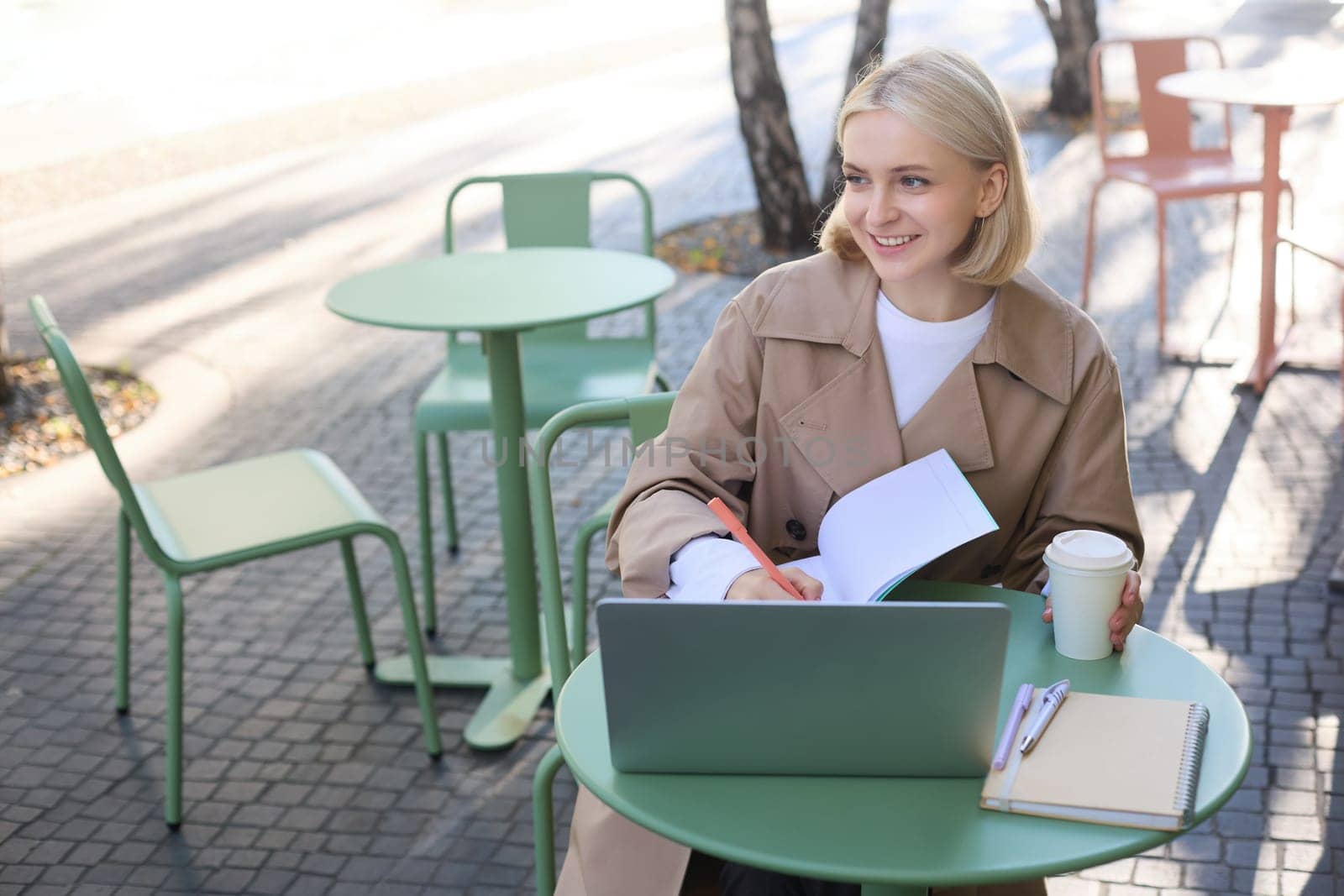 Portrait of young smiling, beautiful woman, studying outdoors in cafe, has laptop and coffee on table, writing in journal, doing homework on sunny day by Benzoix