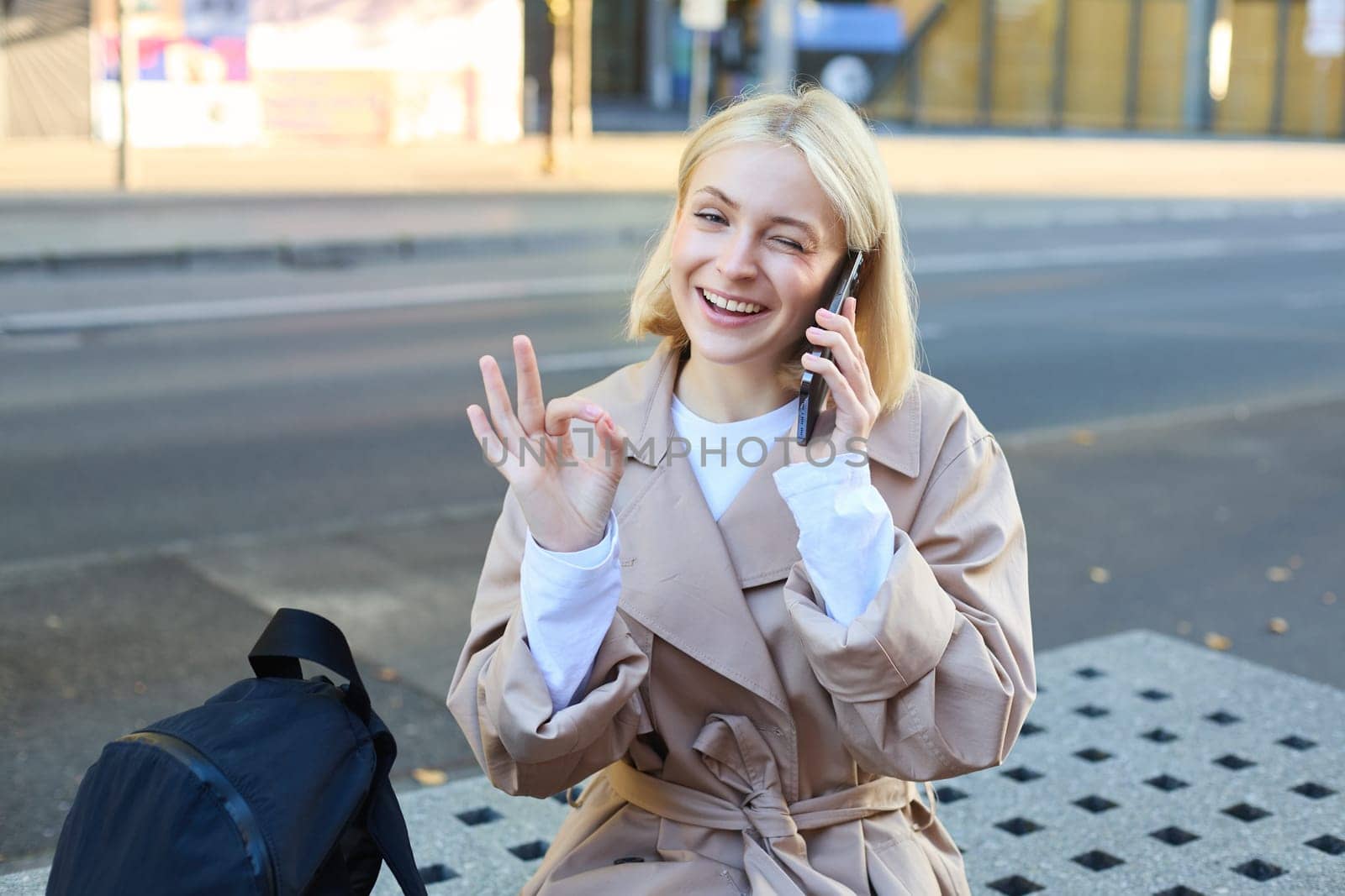 Image of young woman talking on mobile phone, sitting on bench outside, showing okay, ok approval gesture, made a deal over the telephone by Benzoix