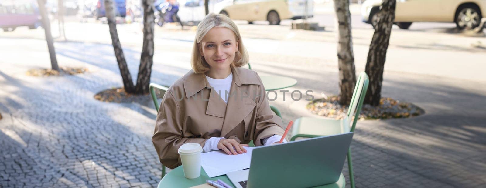 Image of young woman studying outdoors, sitting in cafe, making notes while listening to online lecture using laptop, making notes in notebook, writing.