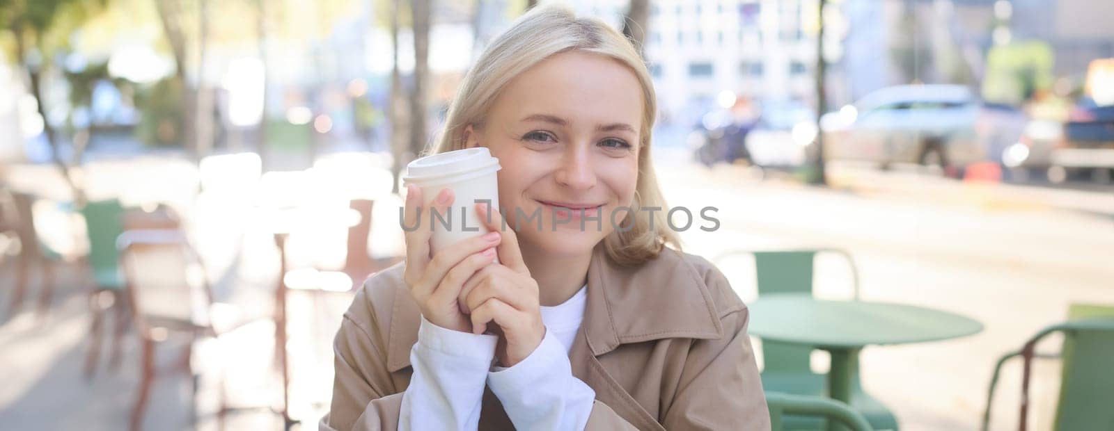 Close up of cheerful blond woman, holding cup of coffee, drinking tea in street outdoor cafe, smiling with happy face expression by Benzoix