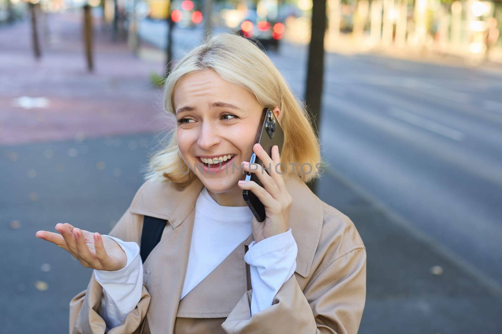 Lifestyle portrait of young blond woman talking on mobile phone, chatting with someone while walking on street, has conversation on smartphone and laughing by Benzoix