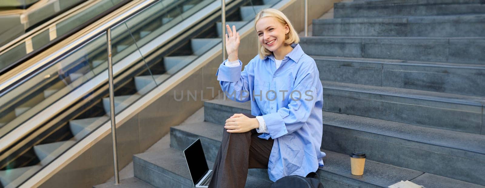 Friendly student girl, saying hi, waving hand at someone, sitting on stairs and drinking coffee, using laptop computer and public wifi.