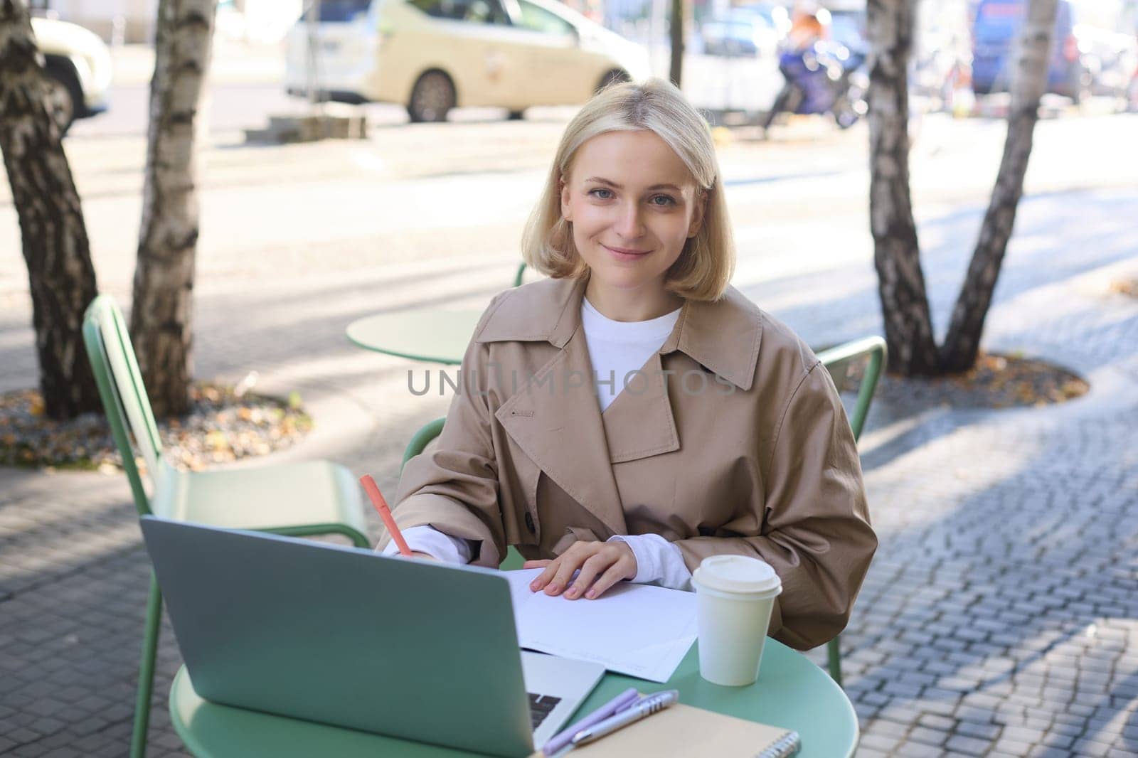 Image of young woman studying outdoors, sitting in cafe, making notes while listening to online lecture using laptop, making notes in notebook, writing by Benzoix